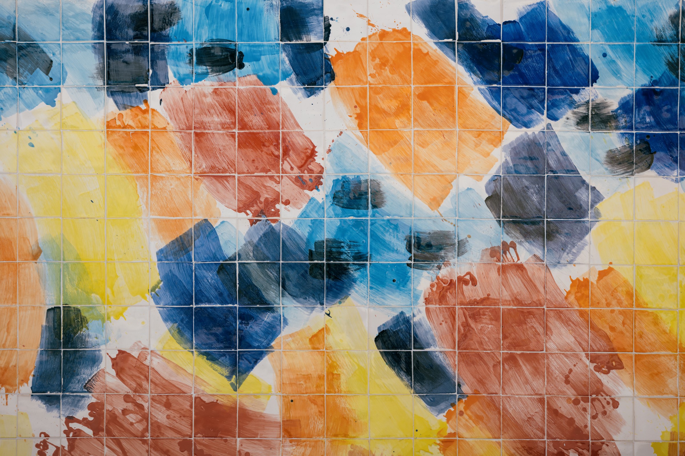 Pormenor de uma superfície revestida por pequenos azulejos quadrados, pintados com pinceladas gestuais em azul, amarelo, laranja, castanho-avermelhado e preto. As manchas de cor sobrepõem-se e distribuem-se de forma irregular, formando uma composição abstrata.