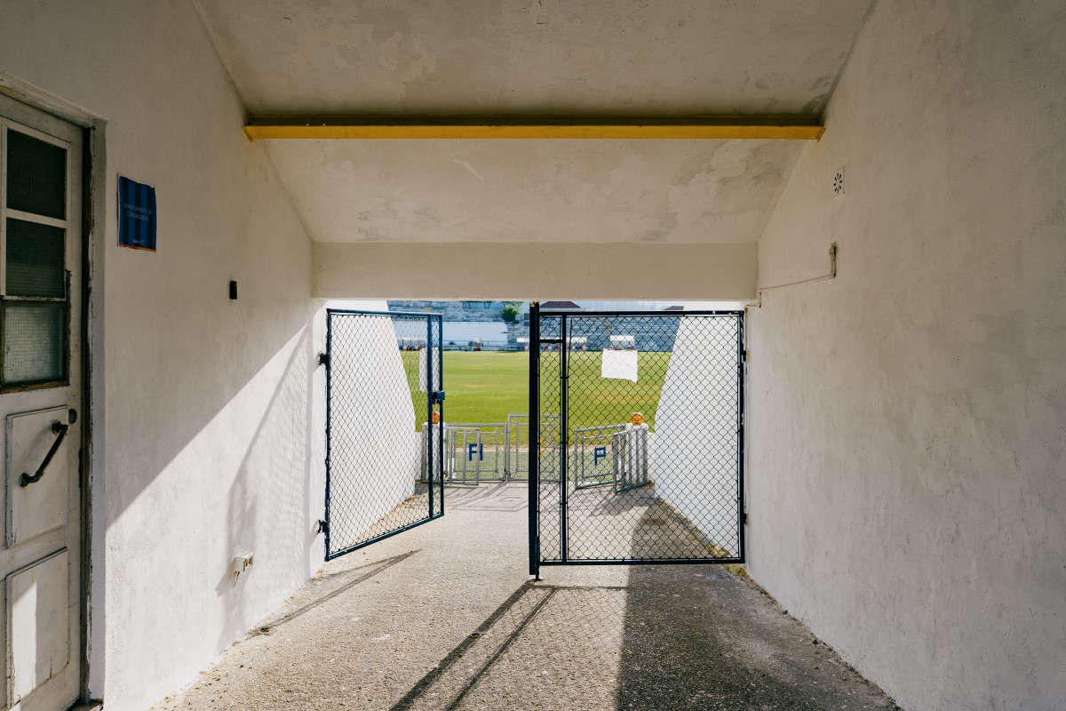 View from the access tunnel leading to the pitch at Tapadinha Stadium. The corridor, with its white walls, leads to an open metal gate through which the football field and part of the stands can be seen in the background.