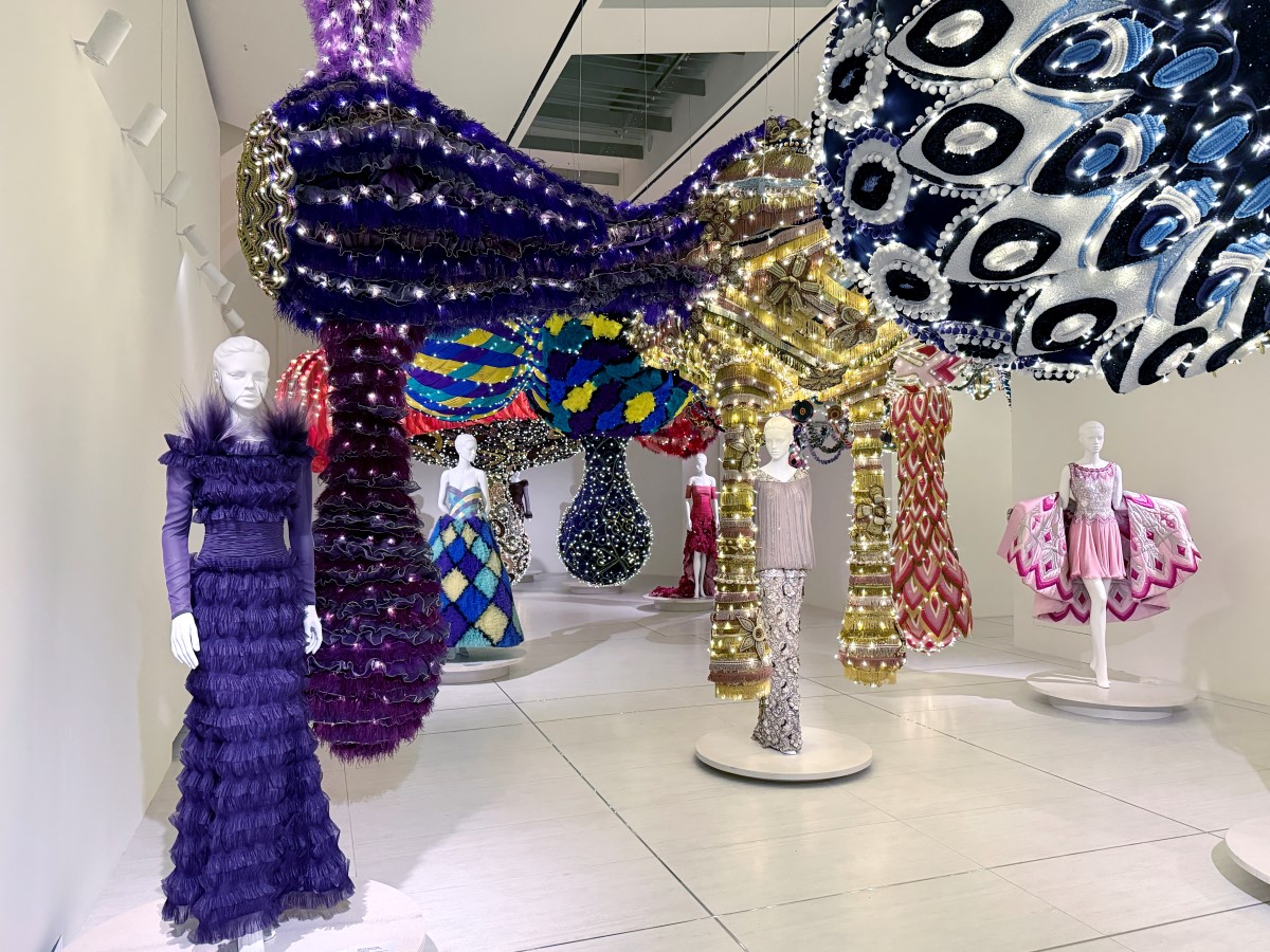 Esculturas têxteis monumentais suspensas entre manequins com vestidos de alta-costura numa galeria expositiva, criando um diálogo entre arte contemporânea e moda.