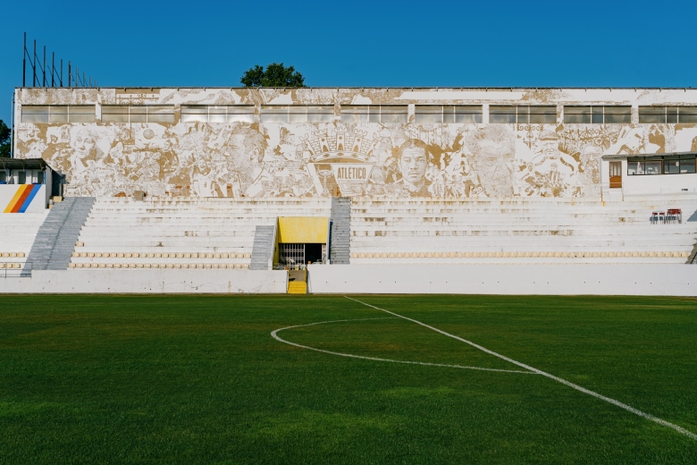 Vista panorâmica do Estádio da Tapadinha, com a intervenção artística de grande escala Scratching the Surface Project, de Vhils, a cobrir toda a fachada da bancada principal. A obra integra retratos, cenas associadas à história do Atlético Clube de Portugal e o emblema do clube ao centro, observados a partir do relvado do estádio.