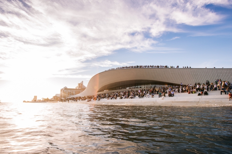 Vista exterior do edifício MAAT junto ao rio Tejo ao pôr do sol, com numerosas pessoas reunidas na cobertura e nas escadarias junto à água.