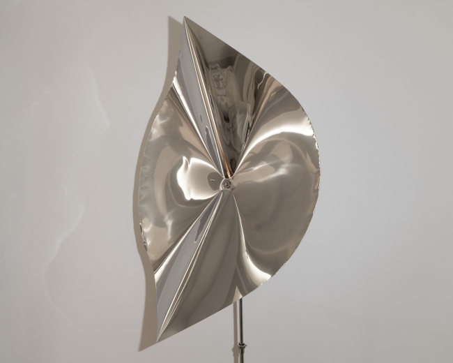 Abstract polished metal sculpture with curved forms and a reflective surface, displayed on a stand against a white wall.