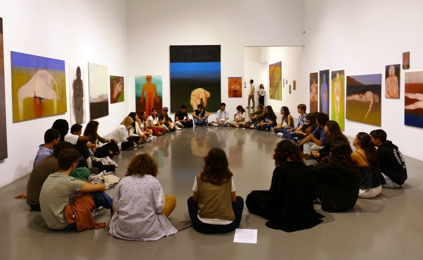 Grupo de jovens sentado em círculo no interior de uma galeria de arte, participando numa atividade de mediação rodeada por pinturas figurativas de grande formato expostas nas paredes.