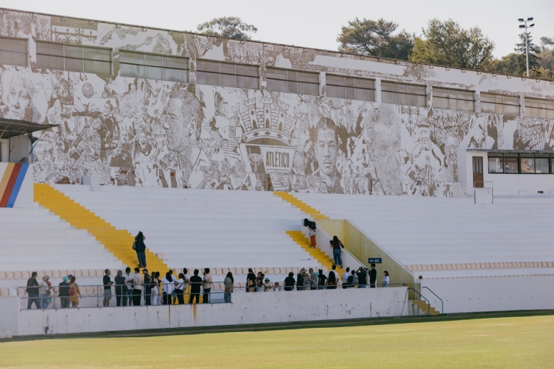 Vista das bancadas do Estádio da Tapadinha com visitantes reunidos junto ao relvado e um mural de grande escala dedicado à história e identidade do Atlético CP.