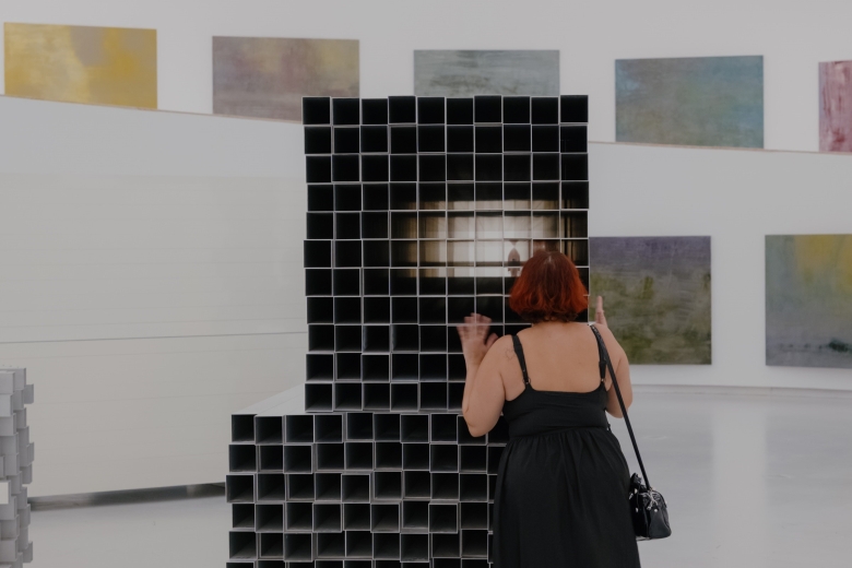 Visitante observa uma escultura geométrica composta por módulos quadrangulares, instalada numa galeria, com pinturas abstratas expostas nas paredes ao fundo.
