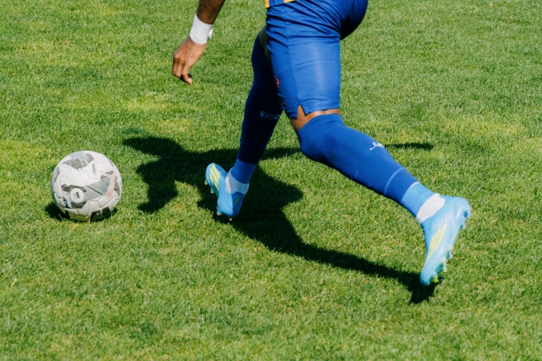 Close-up of a player moving across the pitch at Estádio da Tapadinha, approaching a football. The photograph focuses on the legs, blue football boots and the texture of the grass.