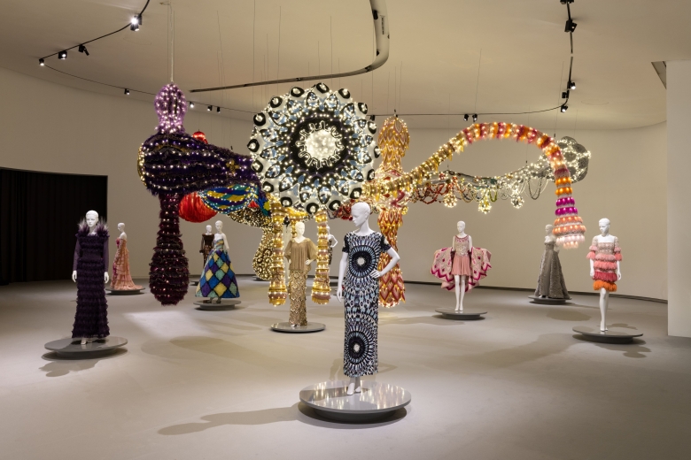 Haute couture garments displayed on mannequins are shown alongside a large suspended installation by Joana Vasconcelos, composed of illuminated, textile and decorative elements within an exhibition gallery.