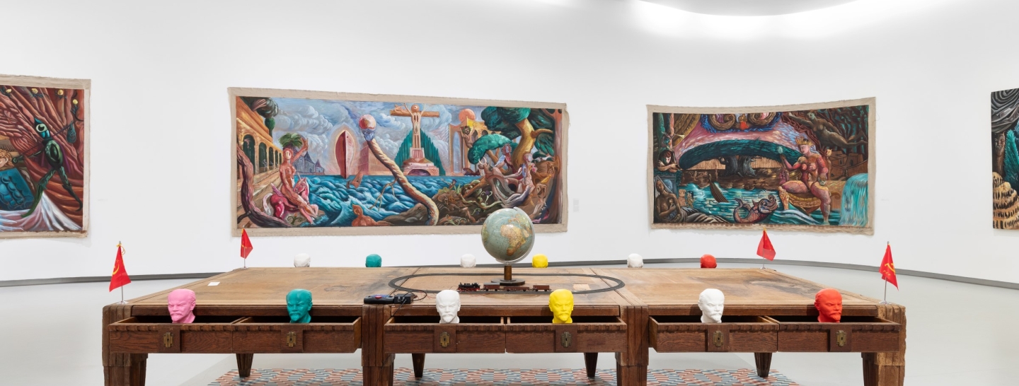 Três mesas de madeira antigas, juntas, com gavetas abertas de onde sai bustos de lenine coloridos; no centro da mesa está um globo com uma linha circular de comboio a circundá-lo; ao fundo, quadros coloridos de Manuel João Vieira